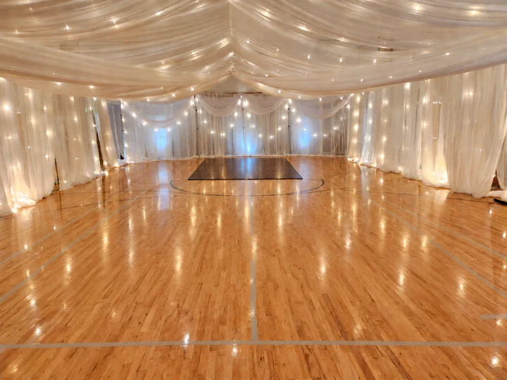 Edited Grand Ballroom room example with canopy, side and end-wall draping, accent swags, string lights, uplights and a dark maple dance floor over the basketball key at the far end.